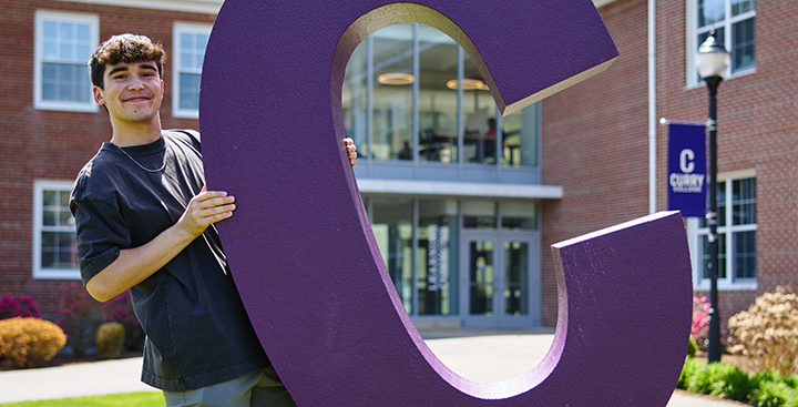A Curry student holds up a giant purple 'C' on the Quad