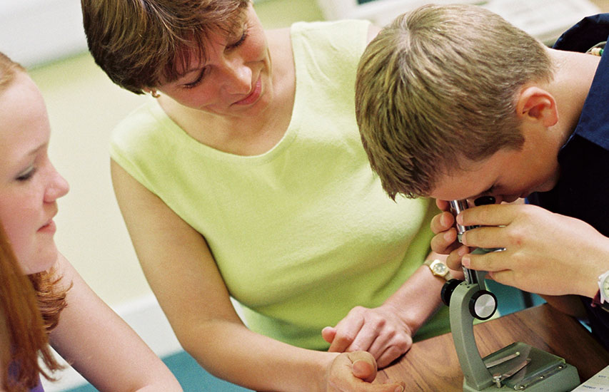 A teacher watches students use a microscopes representing the STEM Education minor at Curry College