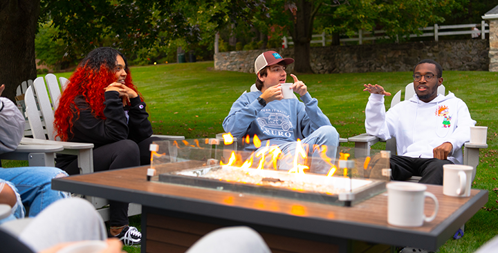 Curry College Student Admission tour guides converse and have hot cocoa at the firepit on campus.